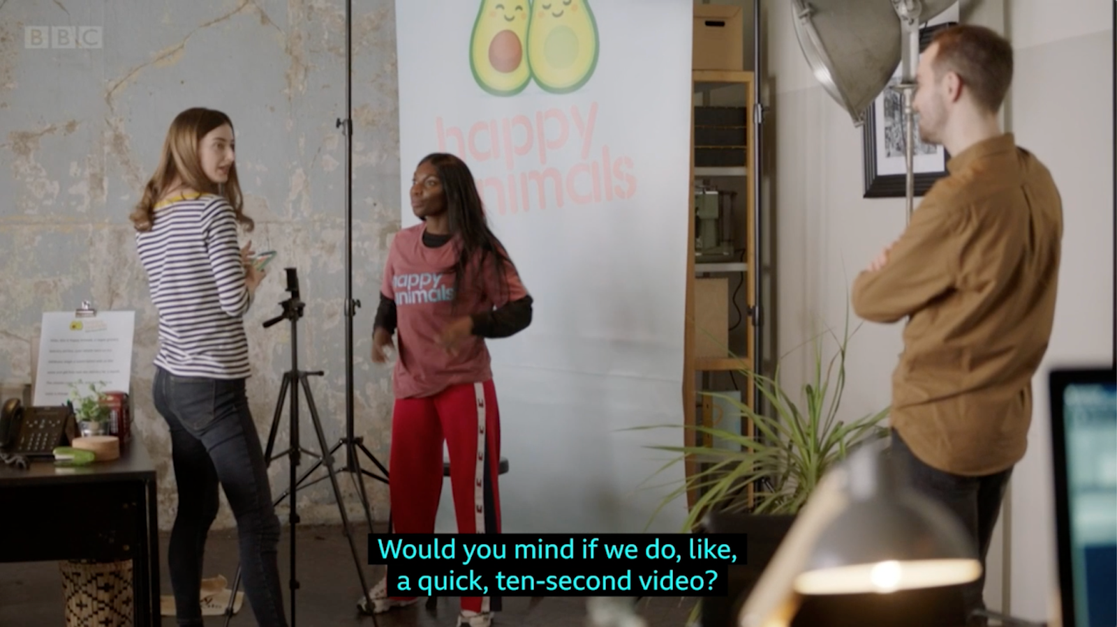 A full shot of Arabella, a slim Black woman with dark long hair, a black top under a pink merch t-shirt that displays the company logo "Happy Animals" and red trousers speaking to Emily, a photographer in front of her. The photographer, a tall, white woman with dark blonde hair, wears a thin striped long sleeve shirt and black jeans, talks to the CEO on her right. The company CEO, a white man with short hair wearing a brown shirt with crossed arms looking to the left to the photographer. On Arabella's right we see a big studio lamp, on her left we see a couple of stands, one of them holding a mobile phone thatis being used to take photos. In the middle, behing Arabella, a long white company banner with the logo consisting two avocado halves next to each other, and below it reads "happy animals" in light pink colour.The subtitles read "Would you mind if we do, like, a quick ten-second video?"