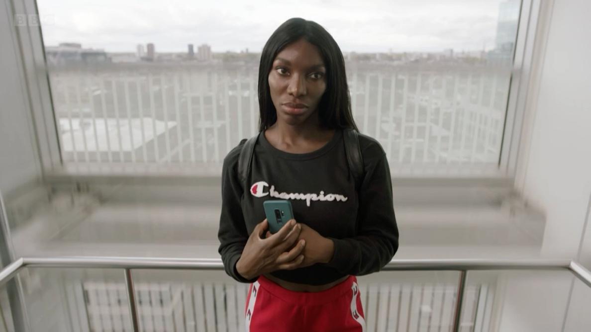 A still image of Arabella, a slim Black woman with dark long hair, on an elevator holding a mobile phone, facing the camera. The woman wears a black top with a Champion logo and red sweat pants. Behind the woman, there is a glass window and a landscape visible through it. The elevator walls are white.