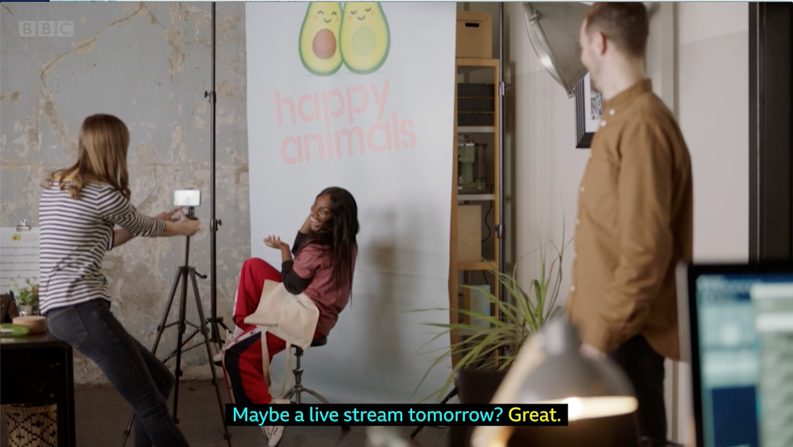 A full shot of Arabella, a slim Black woman with dark long hair, a black top under a pink merch t-shirt that displays the company logo "Happy Animals" and red trousers sitting on a chair, smiling and posing for a photo shoot session. Emily, a photographer in front of her, a white woman with dark blonde hair, wears a thin striped long sleeve shirt and black jeans, while takes a picture of Arabella with a mobile phone on a stand. The company CEO, a white man with short hair wearing a brown shirt with his hands in his pockets looking to the left to the photographer. On Arabella's right we see a big studio lamp, on her left we see a couple of stands, one of them holding a mobile phone thatis being used to take photos. In the middle, behing Arabella, a long white company banner with the logo consisting two avocado halves next to each other, and below it reads "happy animals" in light pink colour.The subtitles read "Maybe a live stream tomorrow? Great"