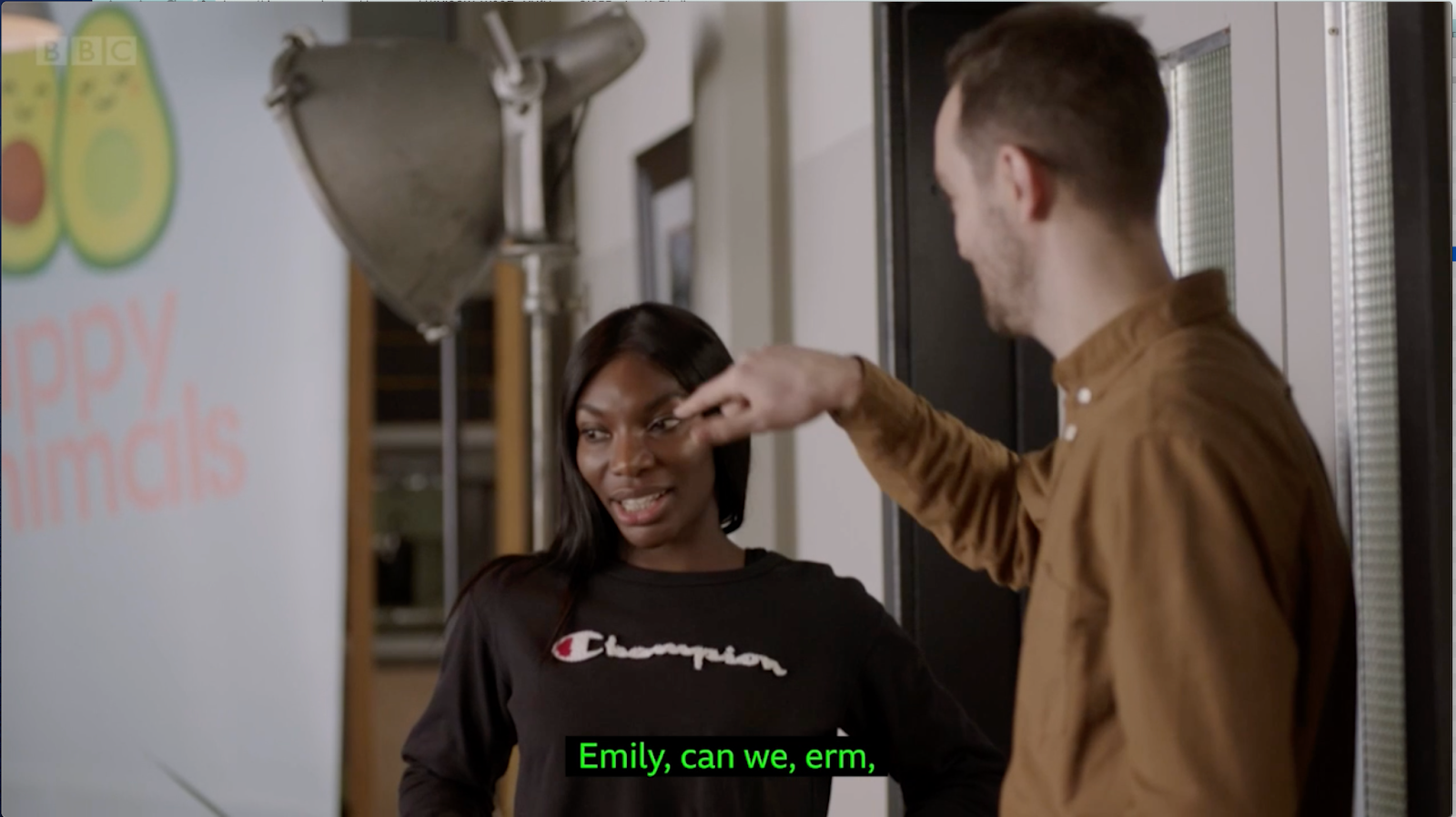 A still image of Arabella, a slim Black woman with dark long hair and a black top talking to the company CEO, a white man with short hair wearing a brown shirt looking and pointing towards the left.  Behind Arabella we see a big studio lamp. On the left, we see part of a white company banner with two avocado halves next to each other, and below it reads "ppy imals" in light pink colour, as the company name is Happy Animals. The subtitles read "Emily, can we, erm,"