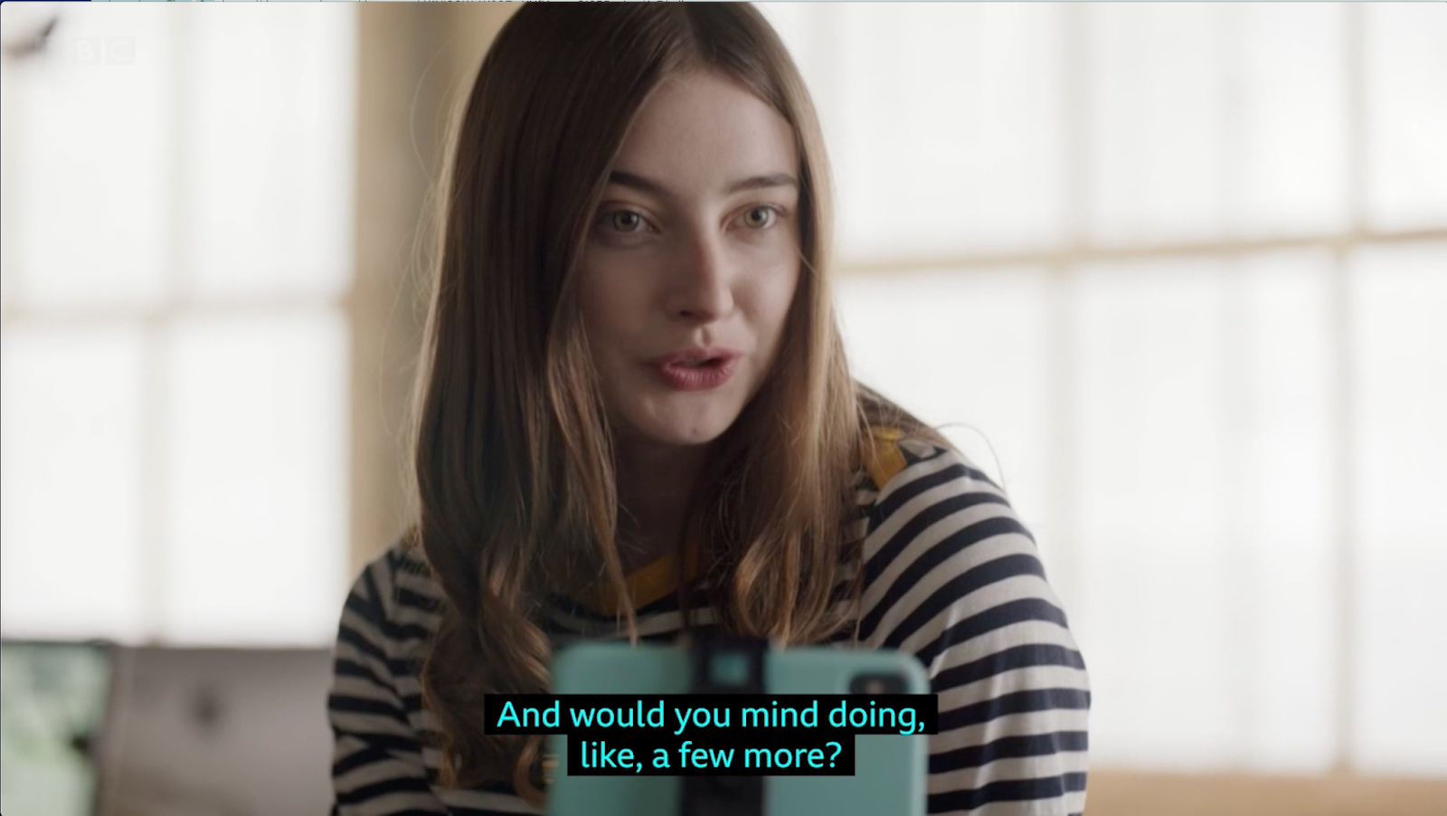 A Mid-close up shot of Emily, the photographer, a white woman with dark blonde hair, wears a thin striped shirt  talking. Behind her, blurry windows with a lot of incoming light. The subtitles read "And would you mind doing like, a few more?"