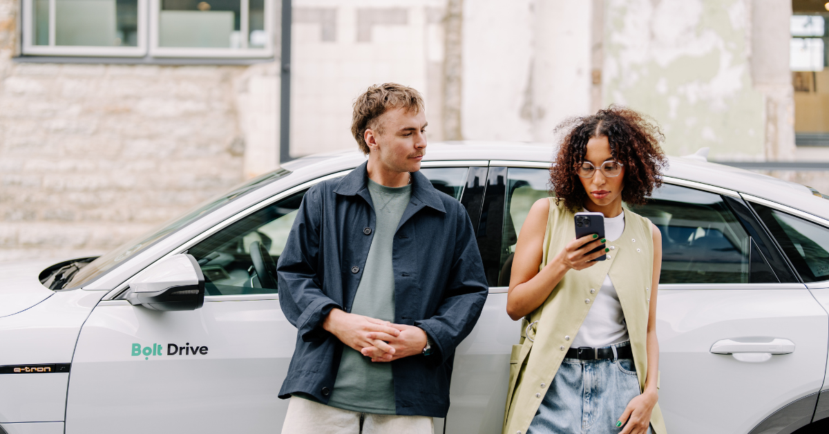 A young man and a woman standing next to a Bolt Drive rental car. The woman is holding a smartphone in her hand.