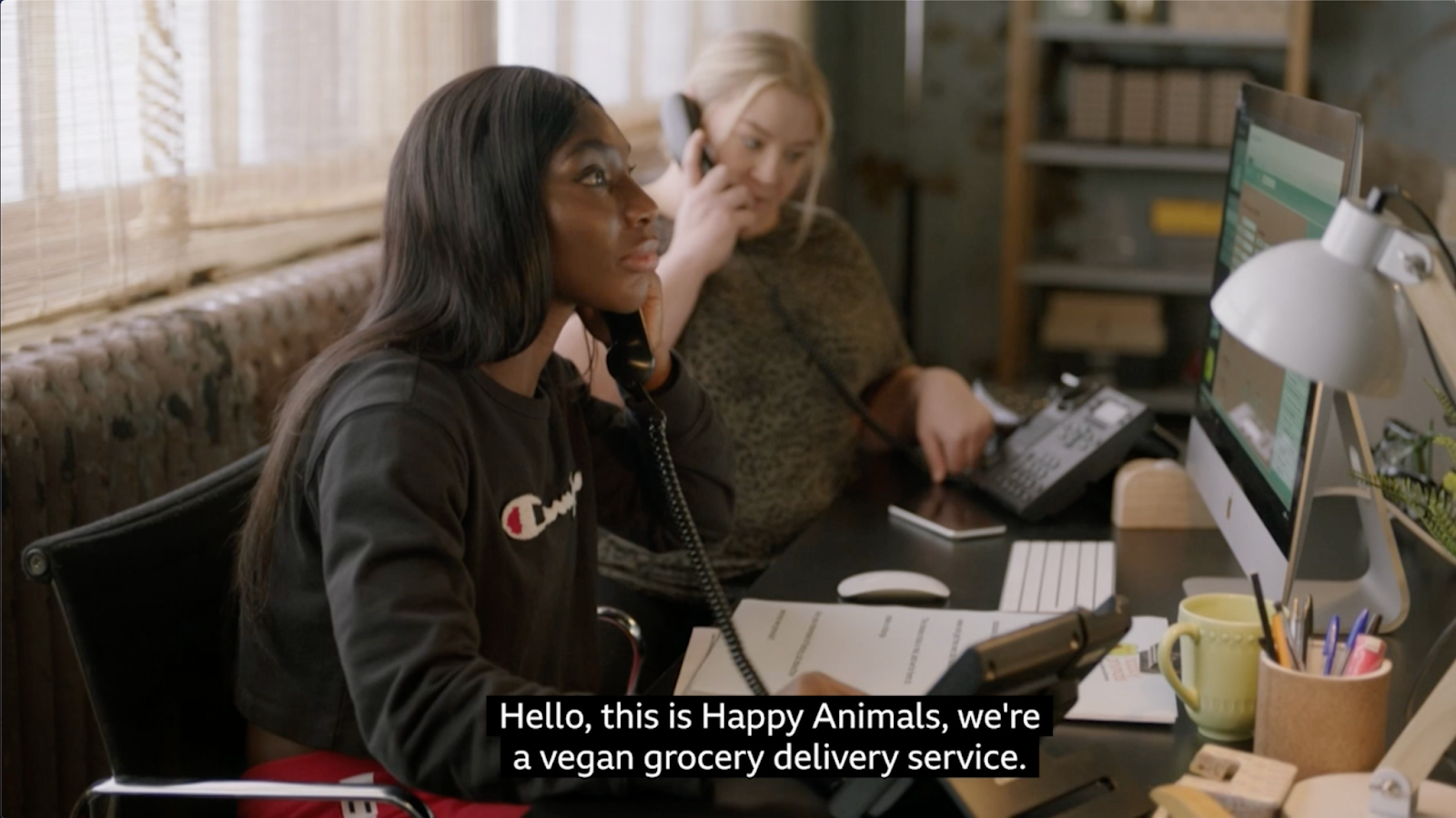 A still image of Arabella, a slim Black woman with dark long hair, sitting on an office chair while answering the phone in front of a computer. The woman is wearing a black top with a Champion logo and red sweat pants. In the background, a white woman sits next to the first woman while also speaking on the phone. The subtitles read Hello, this is Happy Animals, we're a vegan grocery delivery service.