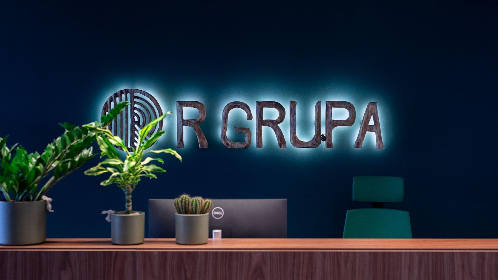R GRUPA logotips uz zilas sienas virs galda uzņēmuma birojā.