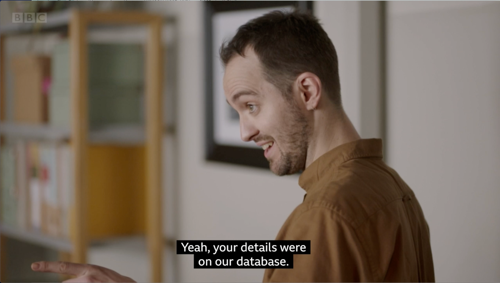 A mid-close up shot of a company CEO, a white man with short hair wearing a brown shirt, looking and pointing towards the left. In the background, we see the white wall and a book shelf with document binders.The subtitles read "Yeah, your details were on our database."